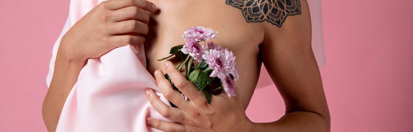 A woman holding pink flowers over her bare chest. She's standing against a pink background and holding a sheet in her other hand.